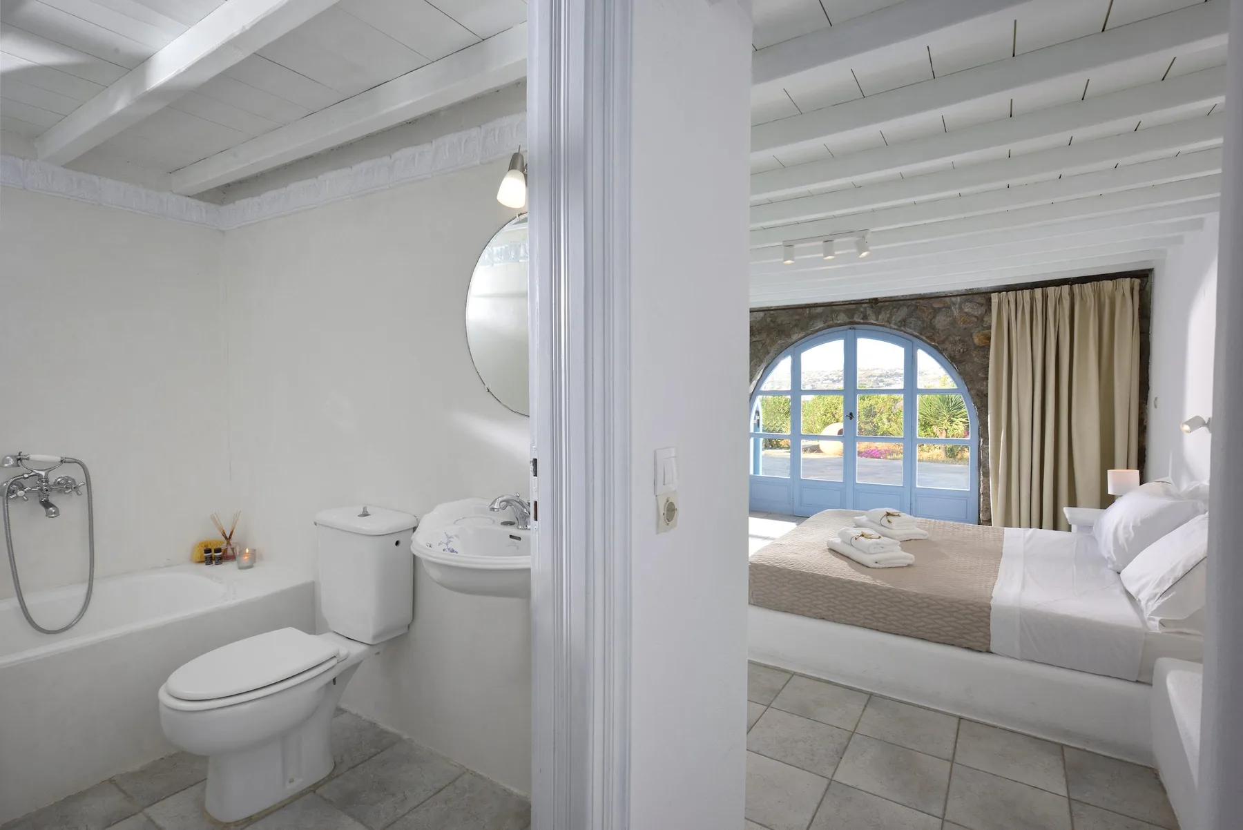 Un dormitorio con una cama blanca junto a una ventana arqueada y un baño contiguo.