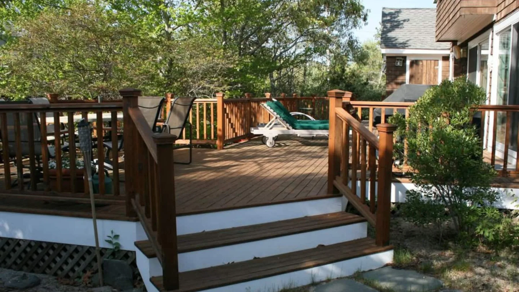 A wooden outdoor deck with steps, railings, and a reclining seat surrounded by trees.