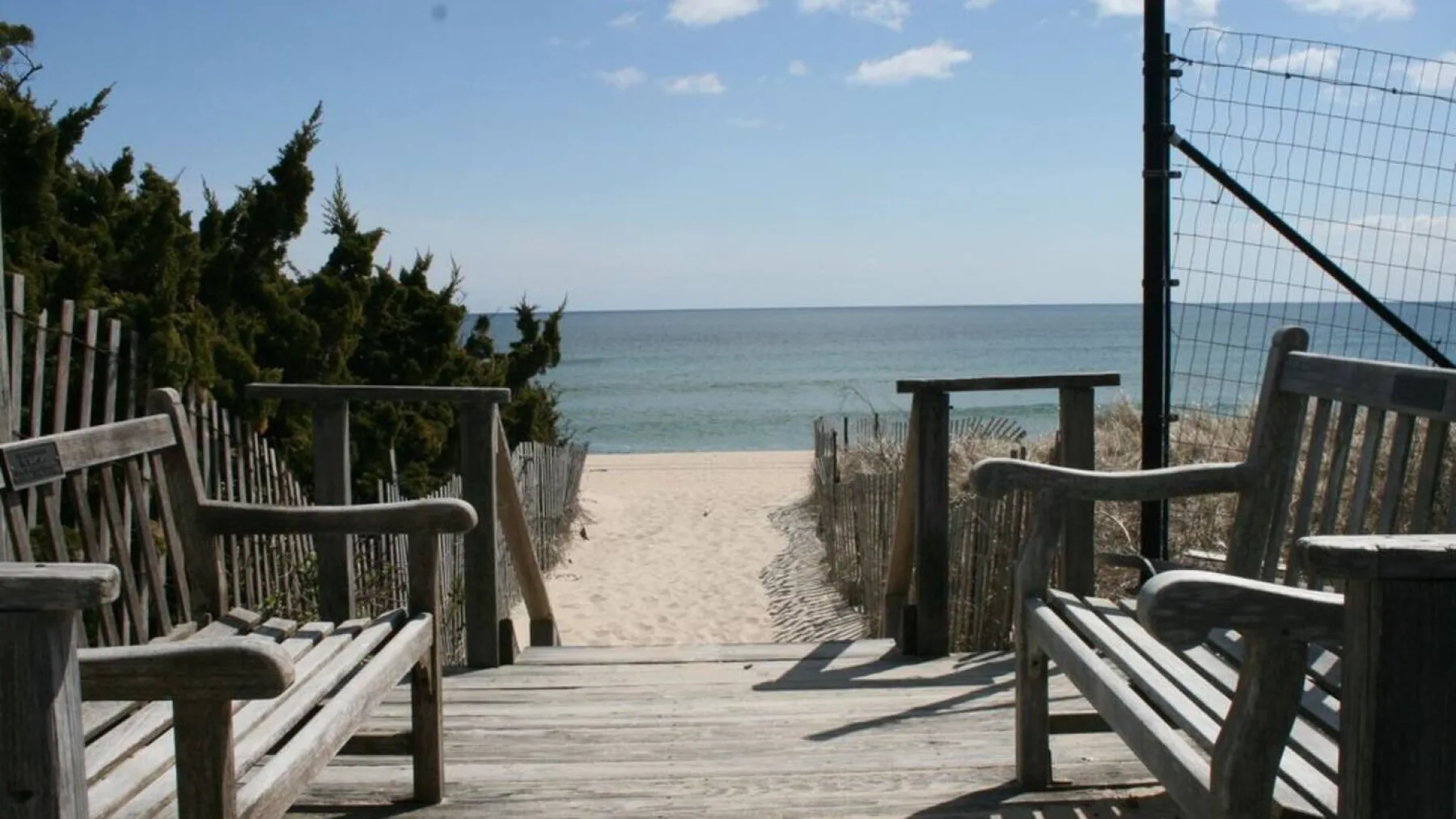 A wooden beach walkway leads between benches toward the sandy shore and ocean.