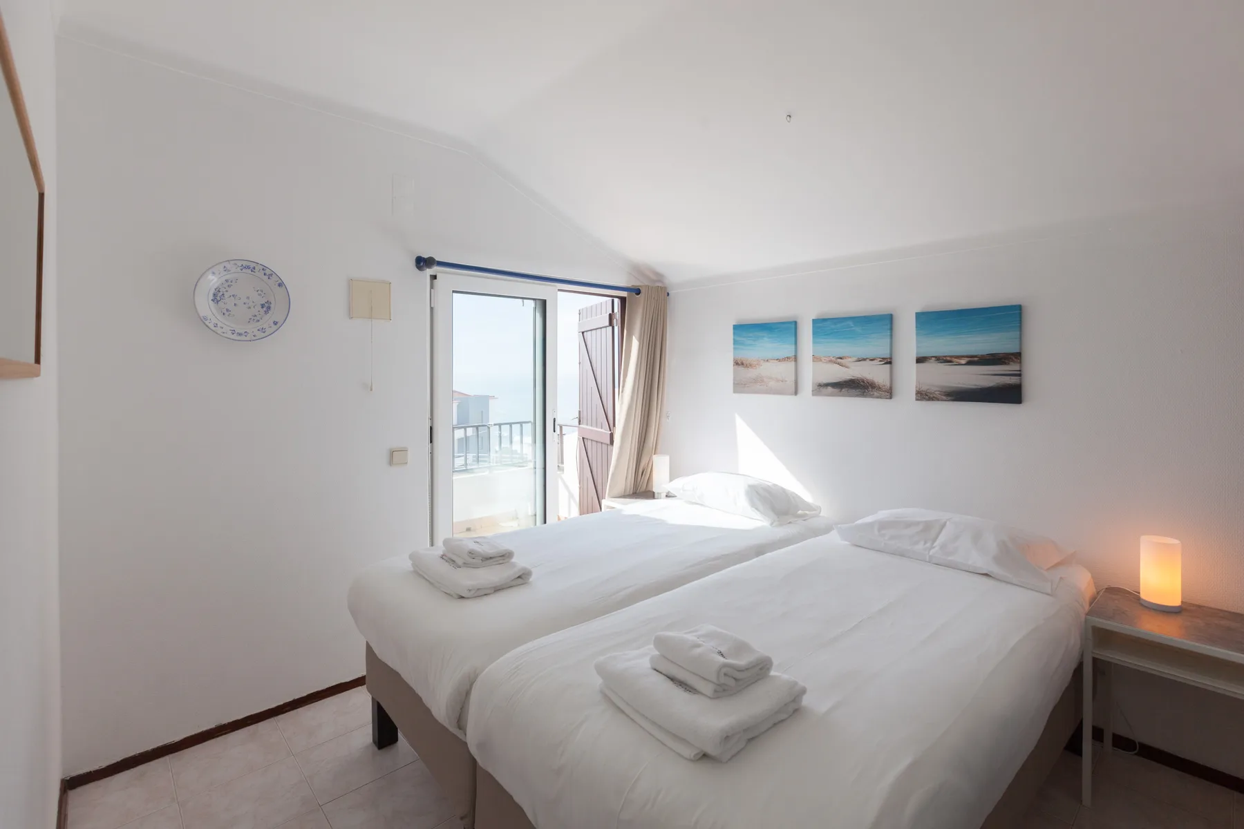A bright bedroom with white bedding and a glass door opening onto a balcony.