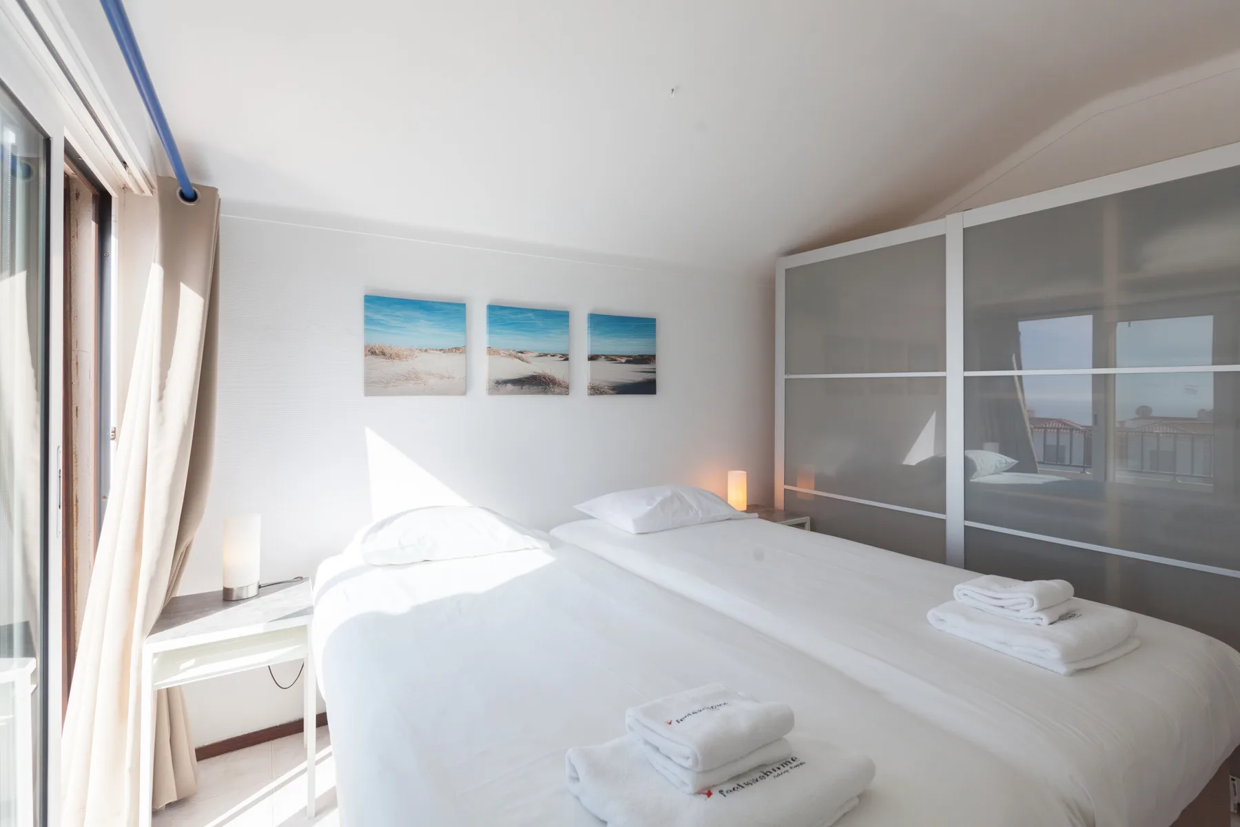 A bright bedroom with a large bed, folded towels, and framed seascapes.