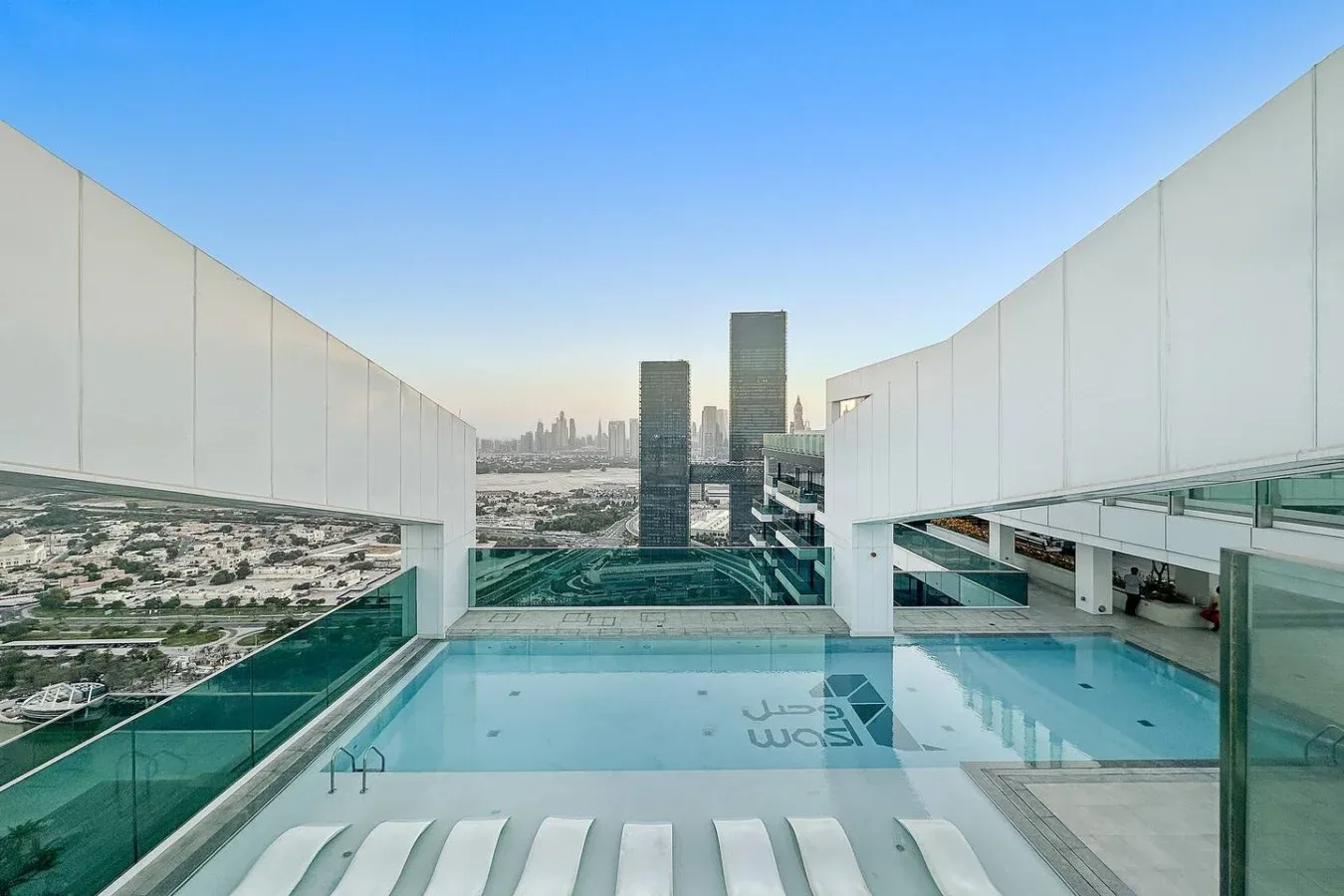 A rooftop pool overlooks a city skyline, with white lounge seating along the edge.
