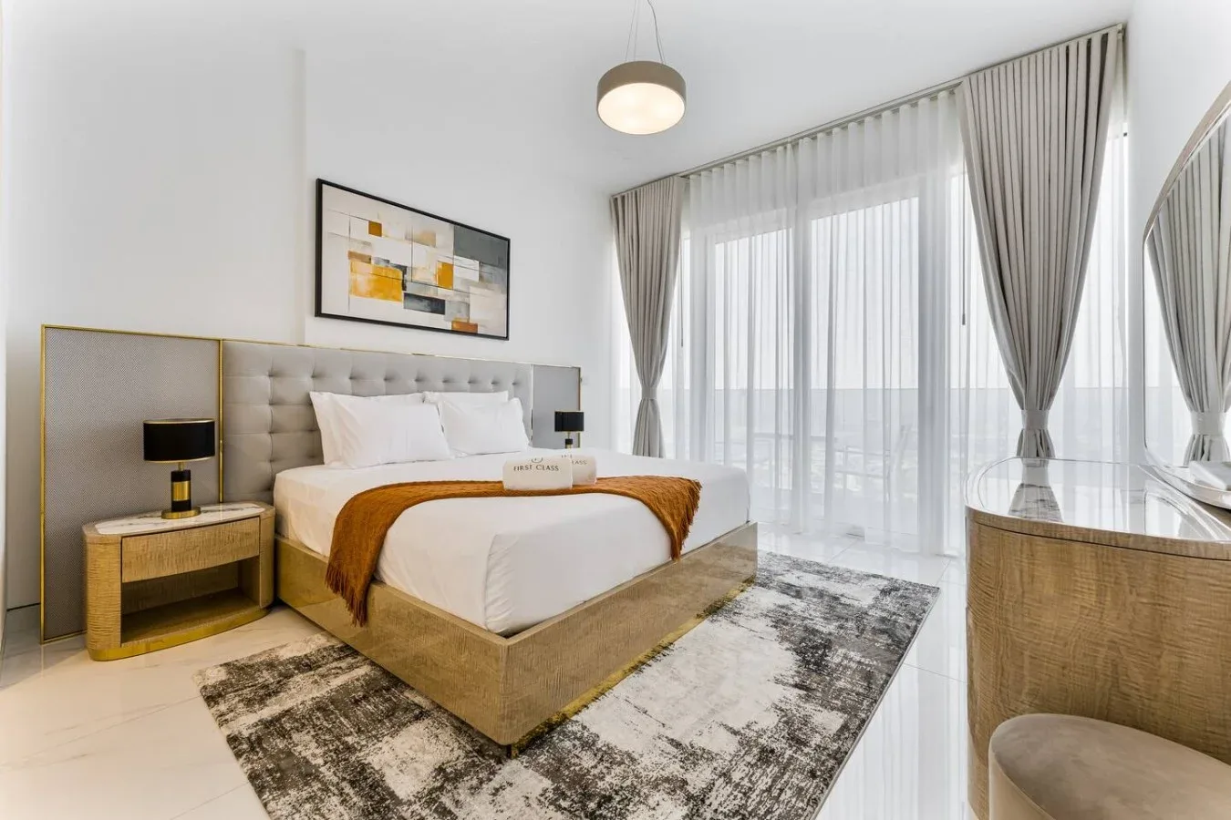 A bright bedroom with a bed, bedside tables, and tall curtained windows.