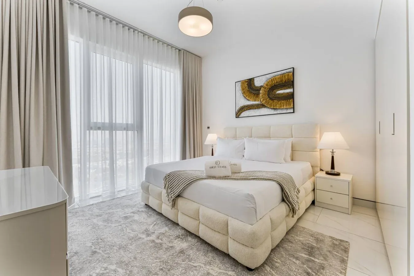 A bright bedroom with a large bed beside a window with sheer curtains.