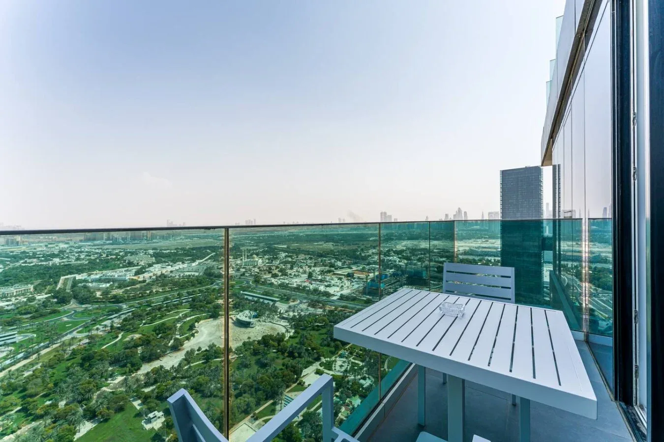 A balcony seating area with a table overlooks a green urban landscape.