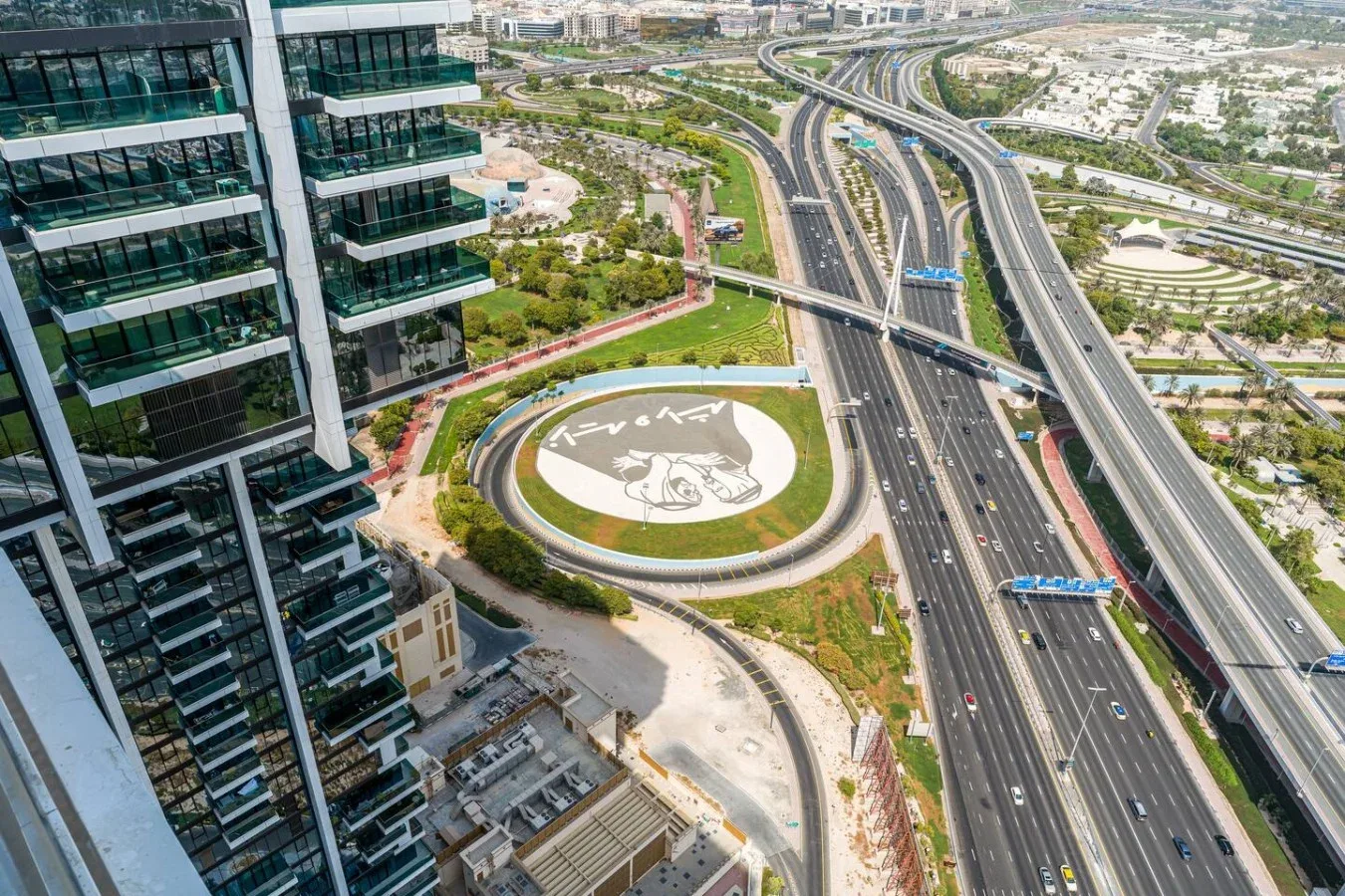 Aerial view of a highway interchange beside modern buildings and a landscaped roundabout.