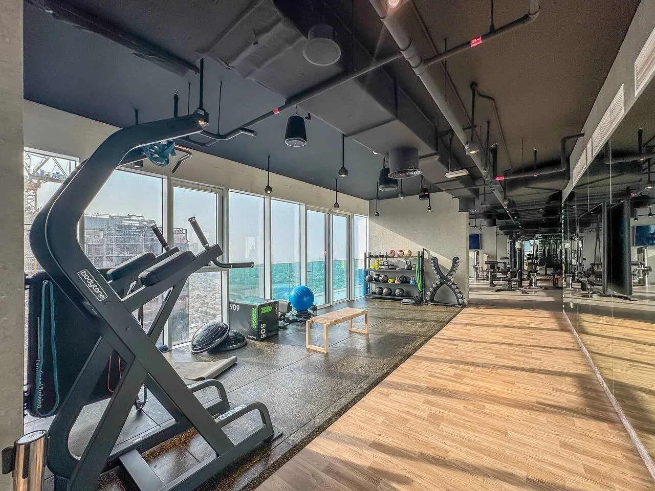 A gym with exercise equipment beside tall windows and a wooden floor.