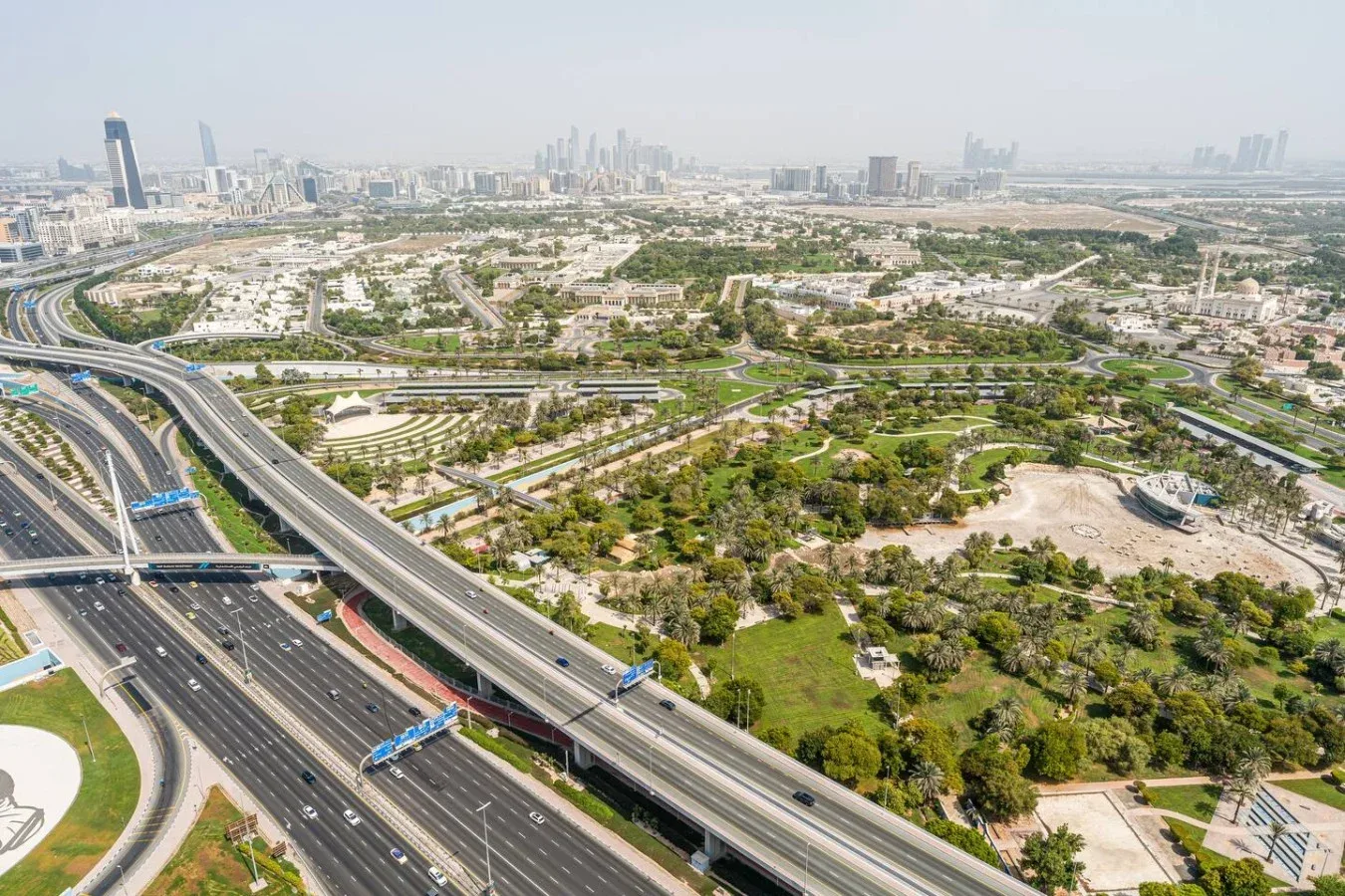 Aerial view of landscaped green spaces and multilane highways, with a city skyline in the distance.