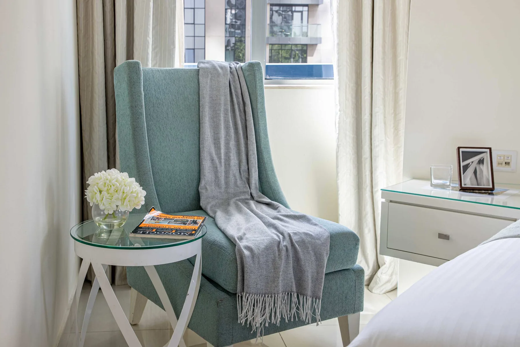 Zona de estar de un dormitorio con sillón verde azulado, mesa auxiliar y ventana.