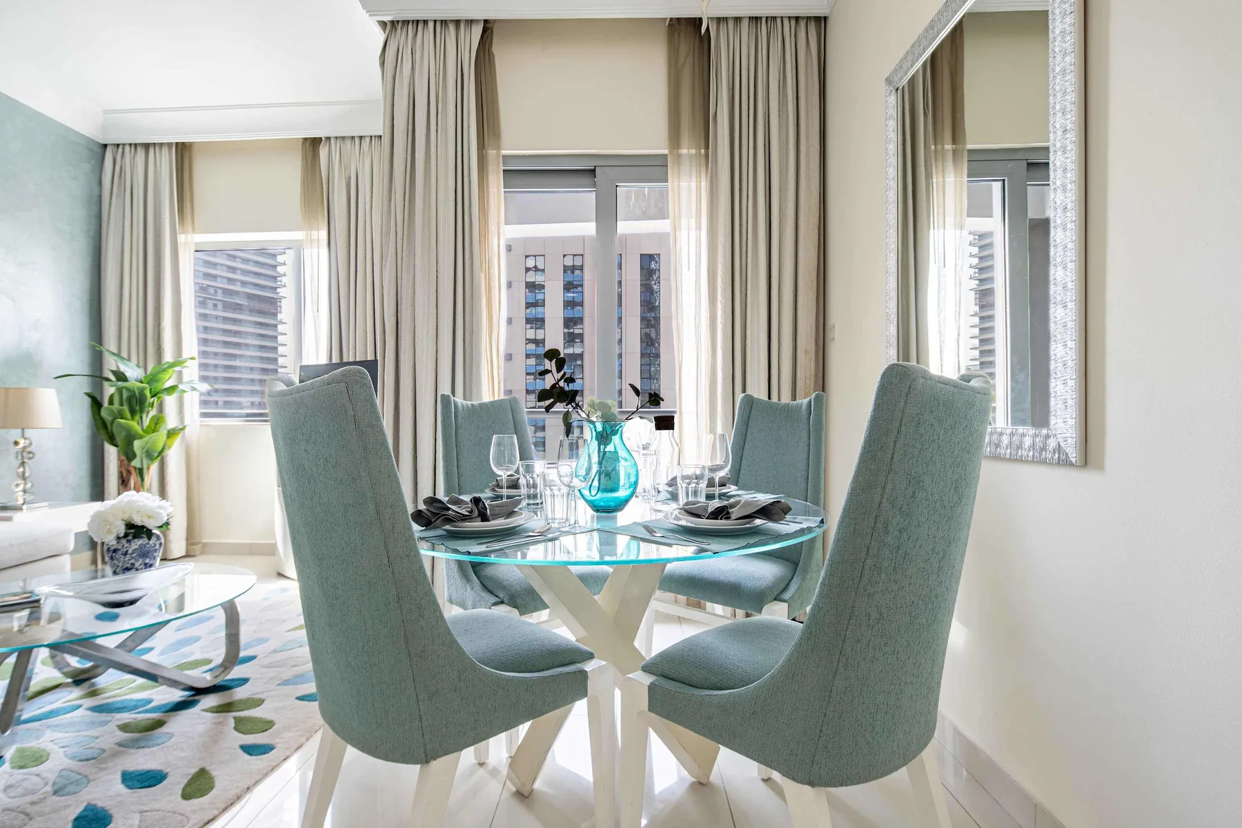 Comedor con asientos tapizados alrededor de una mesa de vidrio junto a ventanas altas.