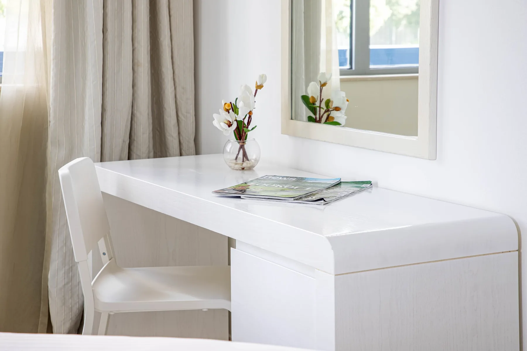 Una habitación luminosa tiene un escritorio blanco, una silla y un espejo.