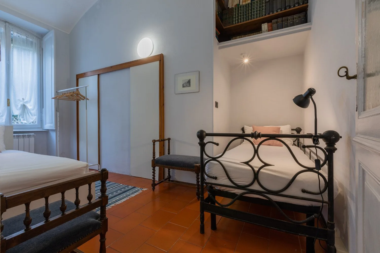 A bedroom with two beds, a tall window, and warm terracotta-colored flooring.