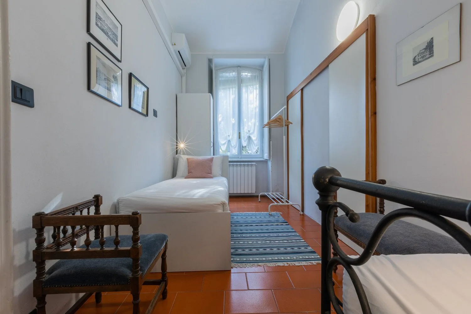 A narrow bedroom with a single bed, a window, and a striped rug.