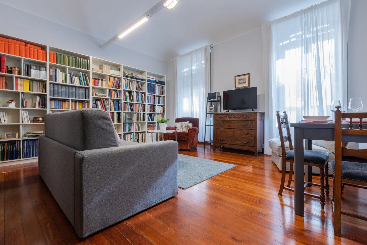 A living room with bookshelves, a sofa, and a dining table beside tall windows.