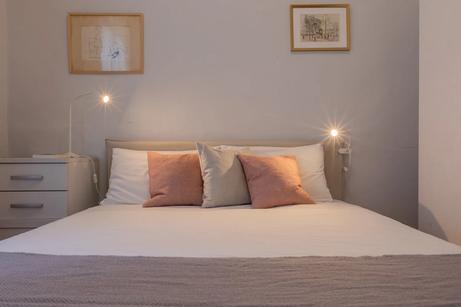 A neatly made bed with pillows and bedside lamps sits against a plain wall.