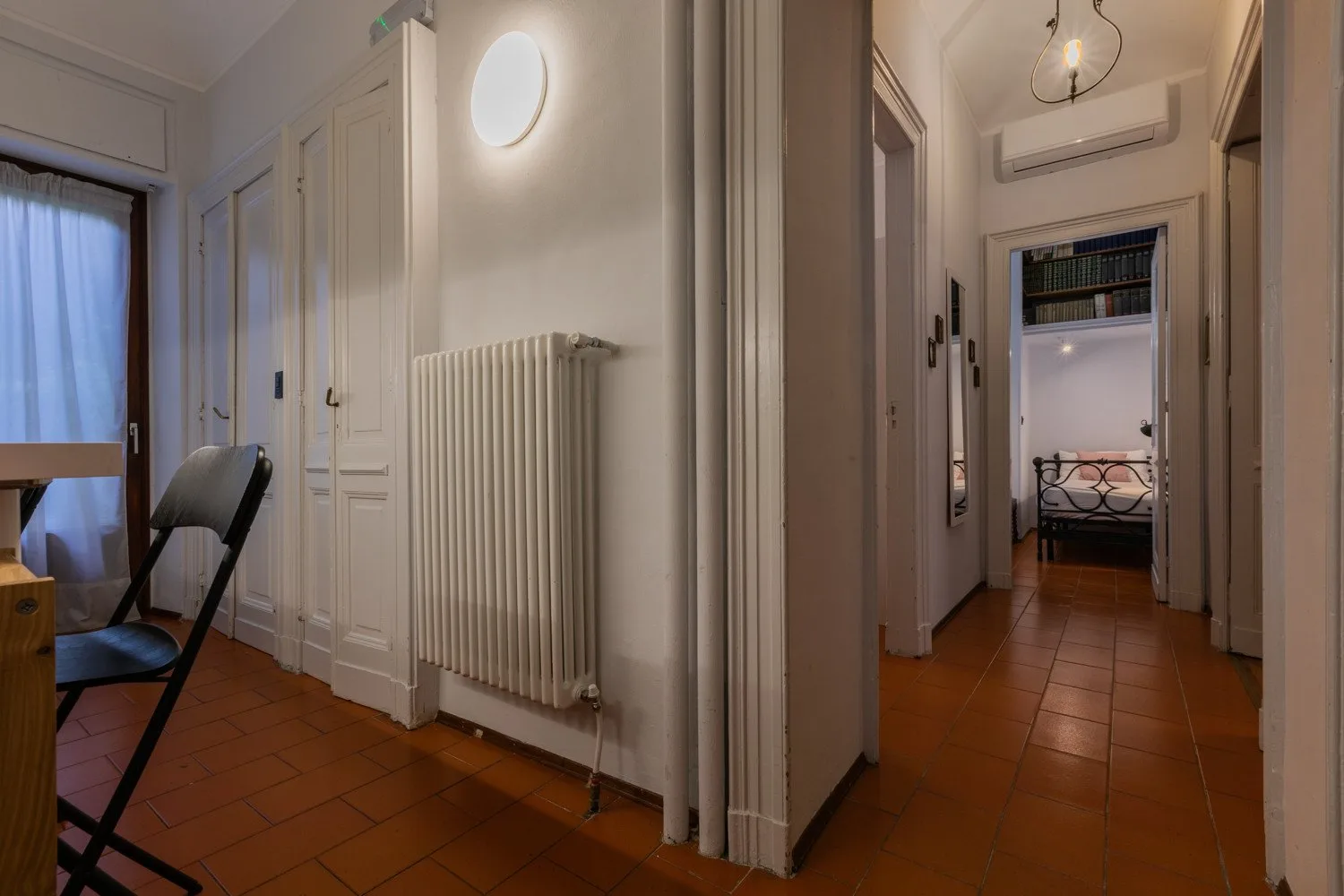 A tiled hallway with doorways leads to a room with a bed.