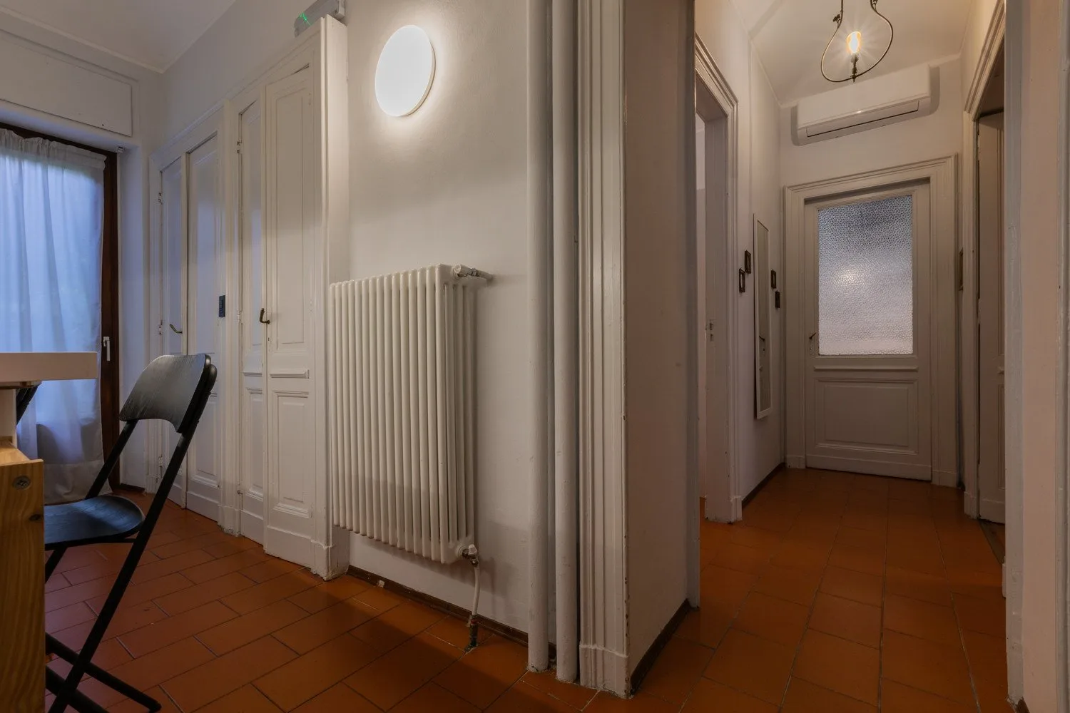 A narrow interior hallway with white doors, a radiator, and terracotta-colored floor tiles.