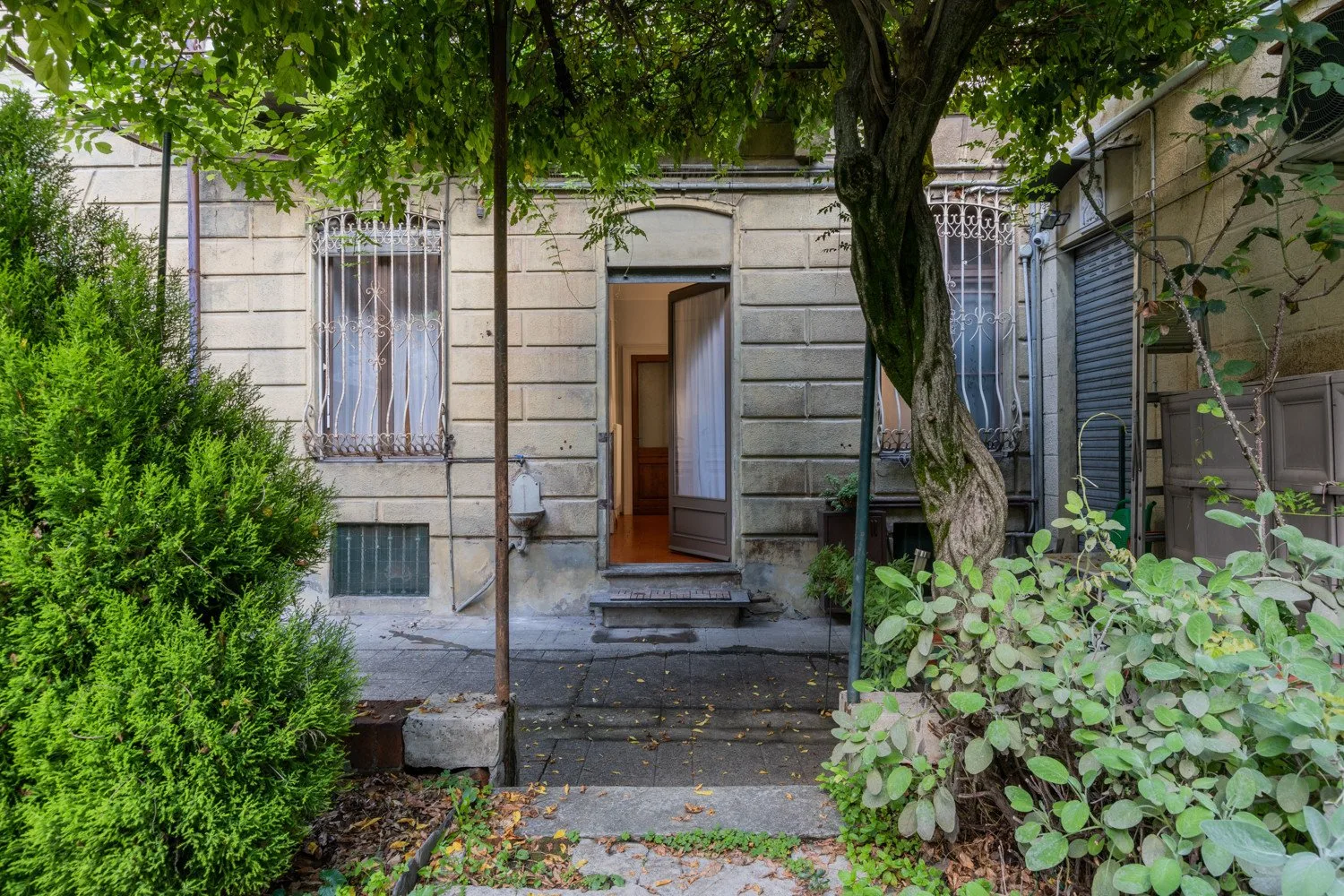 A stone building entrance opens onto a garden path beneath leafy trees.