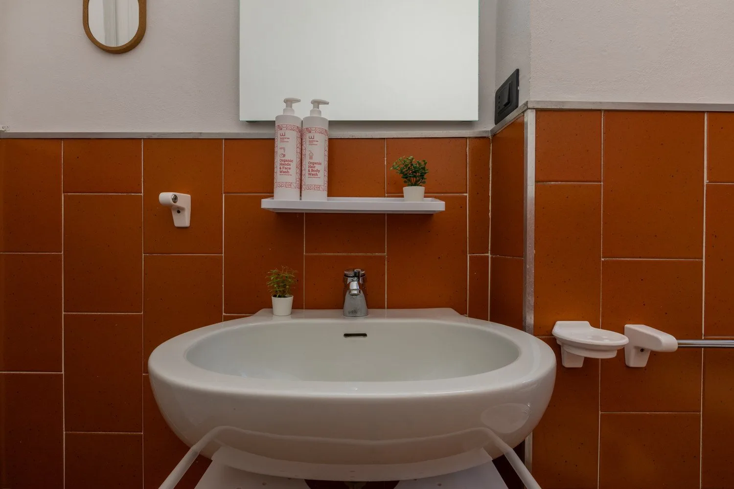 A bathroom sink sits against orange tiled walls beneath a small shelf.