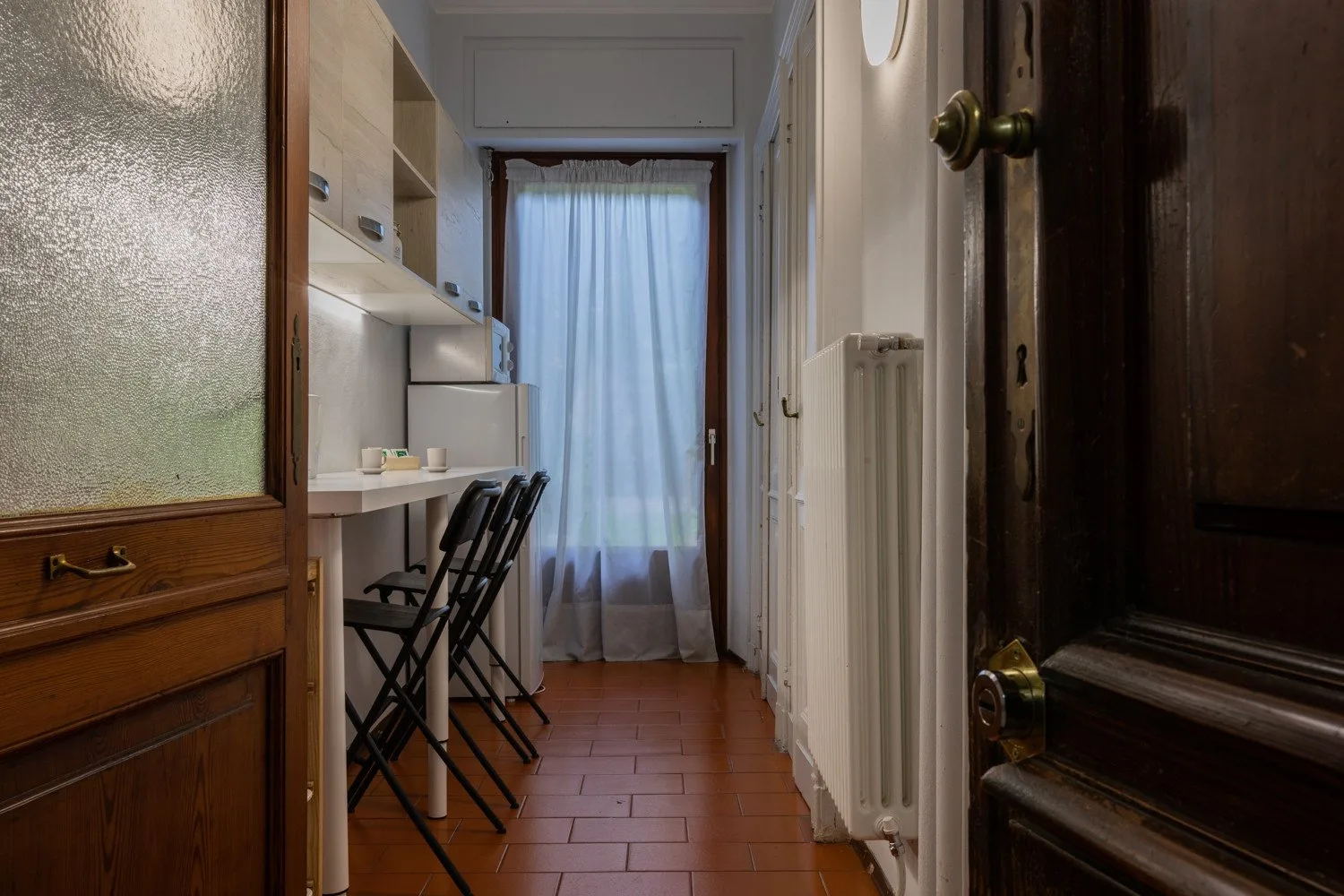A narrow hallway with a small desk and chairs beside a curtained window.