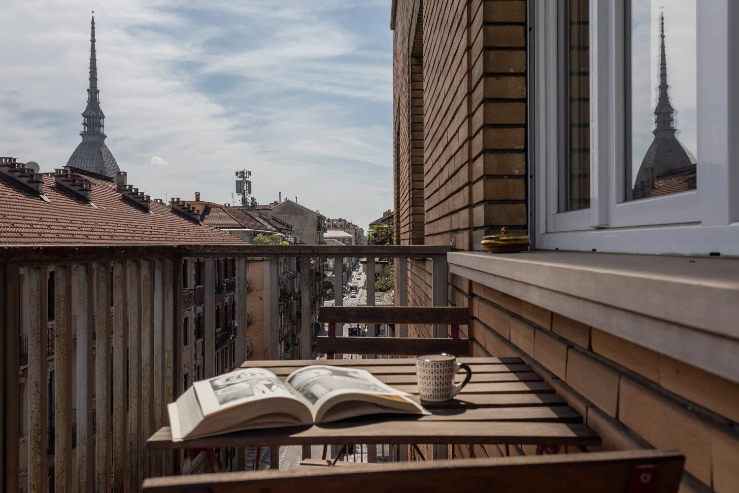 Una zona de estar en un balcón da a los tejados y a una aguja lejana, con un libro abierto sobre la mesa.