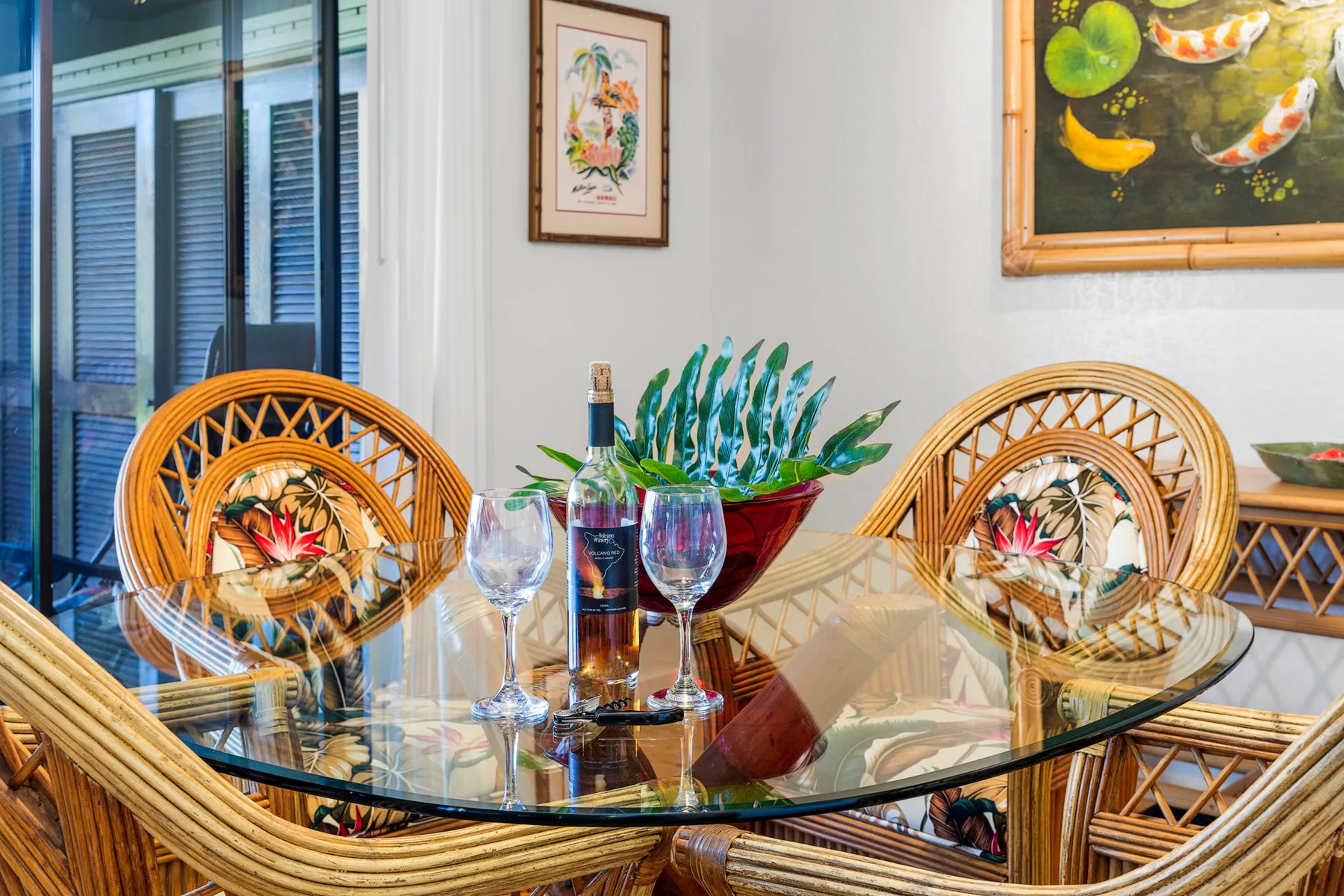 Un comedor con mesa de vidrio, asientos tejidos y cuadros enmarcados.