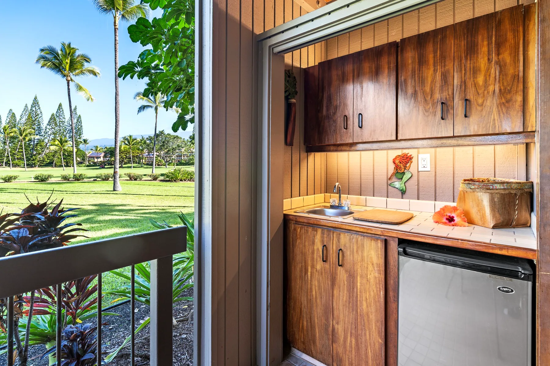 A kitchenette with wooden cabinets and a sink opens onto a green landscaped view.