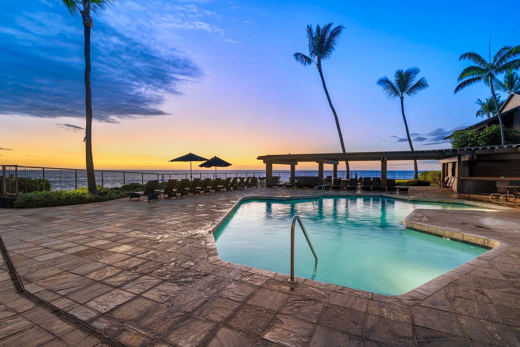 An outdoor swimming pool and paved deck overlook the ocean at sunset.