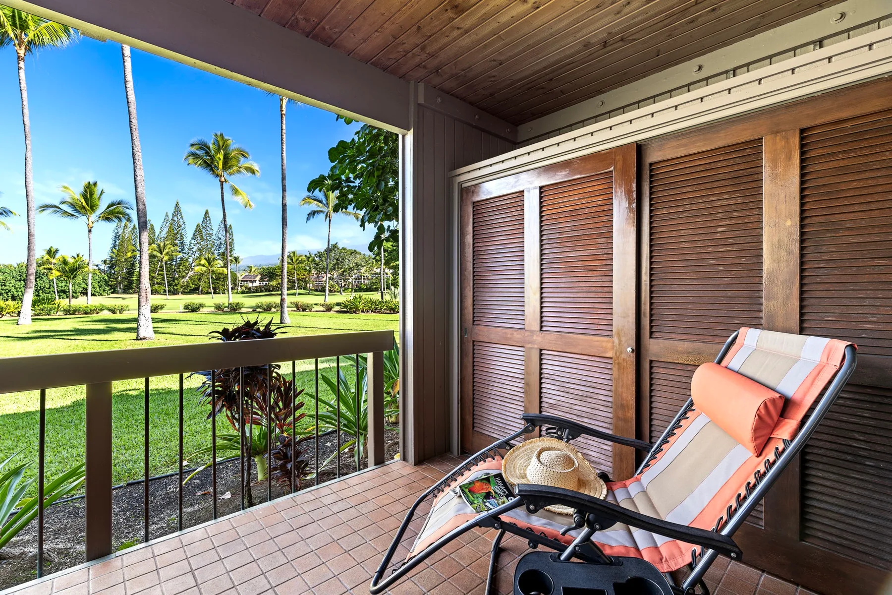 A covered balcony with a reclining seat overlooks a green lawn and palm trees.
