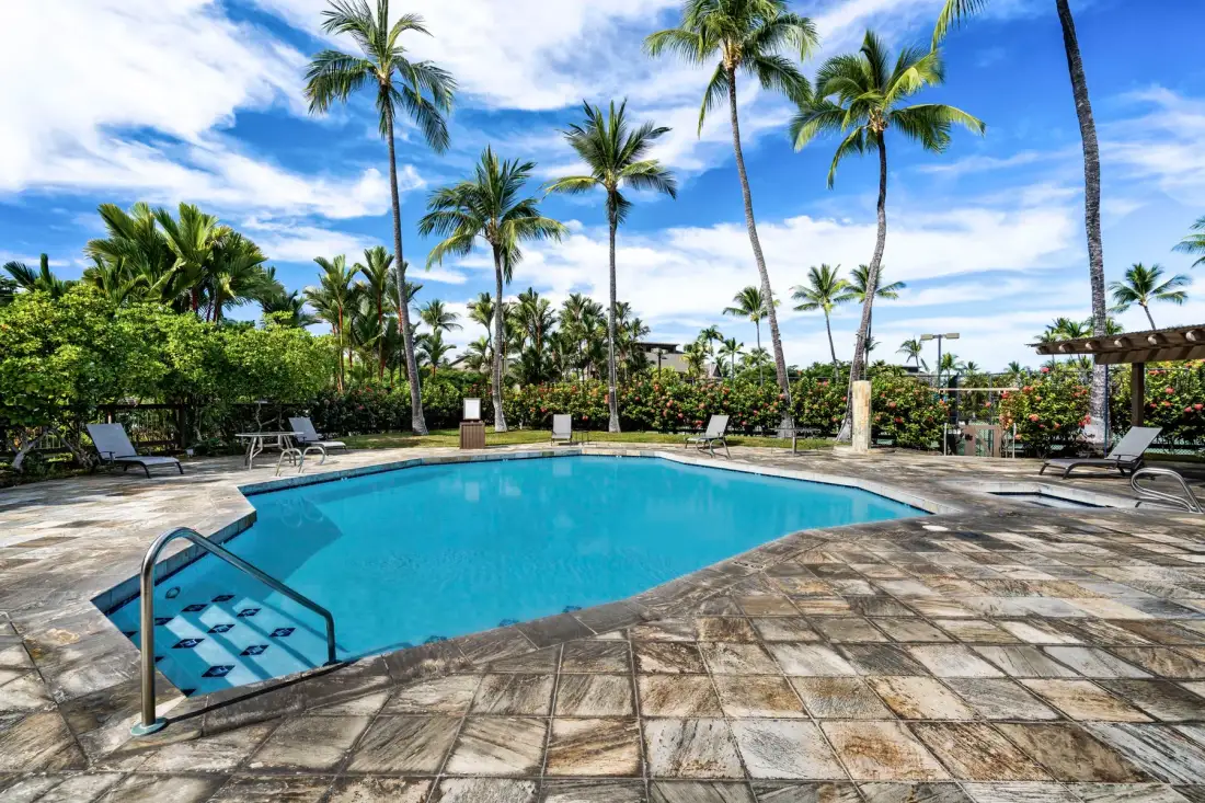 An outdoor swimming pool is surrounded by palm trees and paved seating areas.