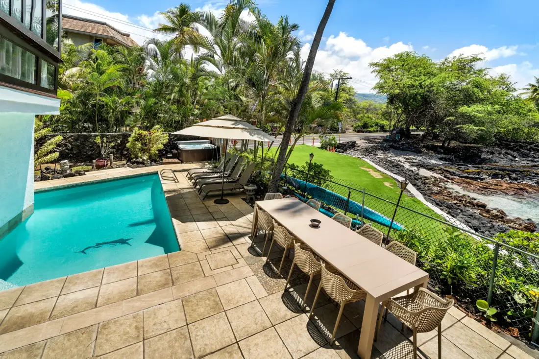 Un patio junto a la piscina con asientos al aire libre da a un jardín verde y una costa rocosa.