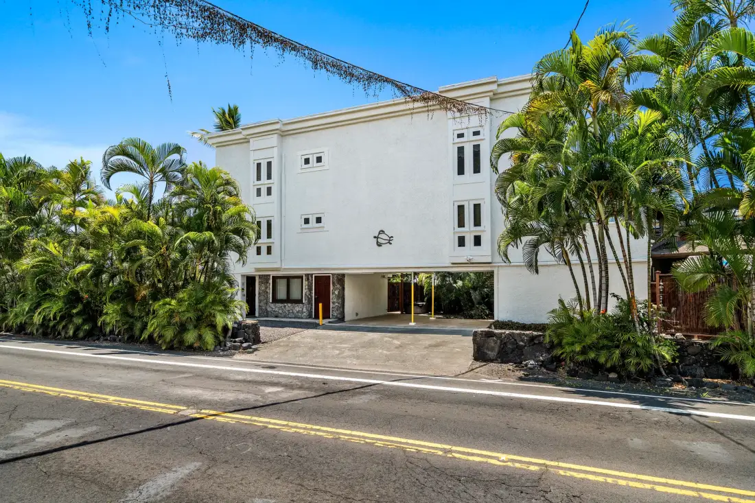 Un edificio blanco de varias plantas se alza tras palmeras tropicales junto a una carretera.