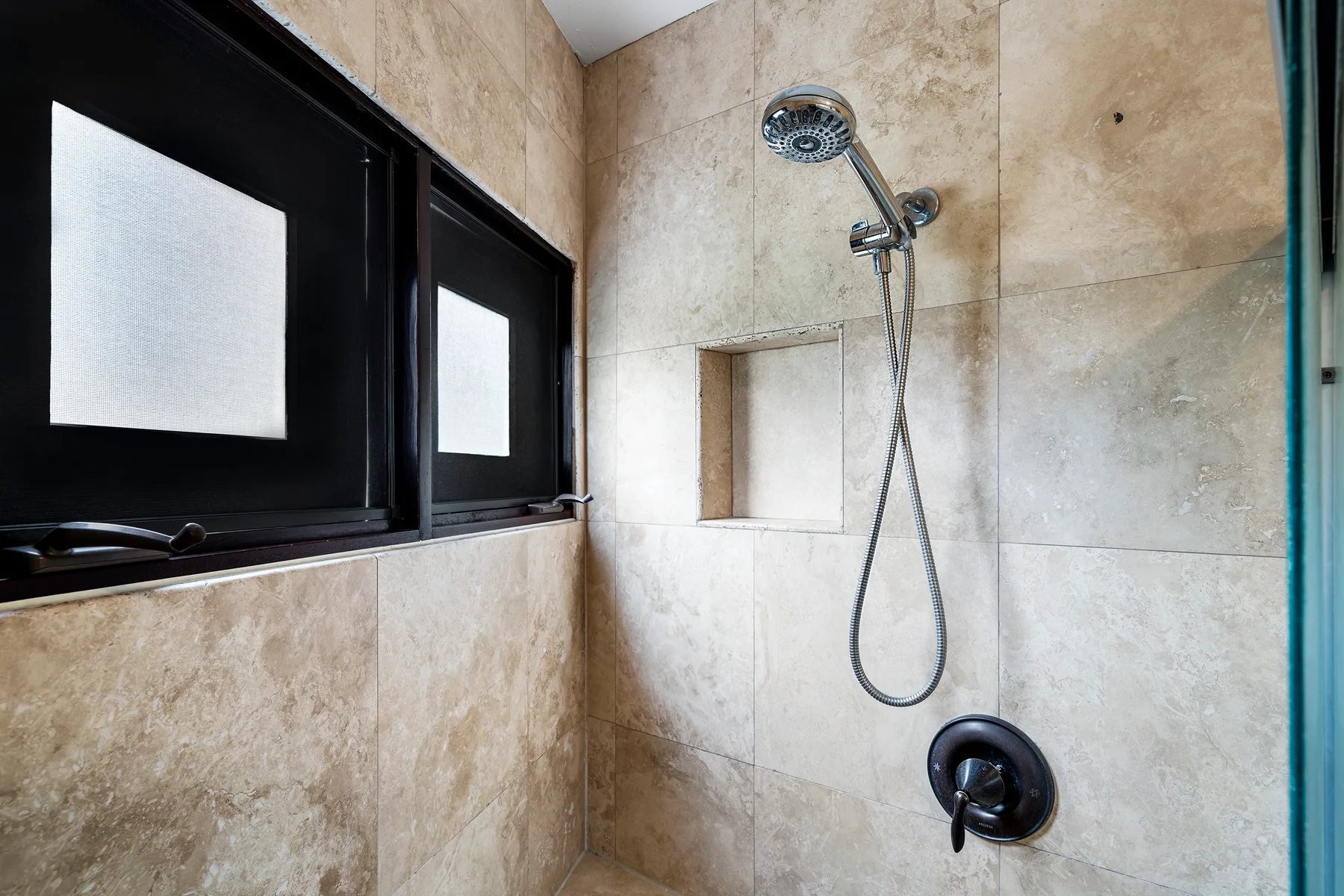 Zona de ducha con azulejos, ducha de mano y una ventana.