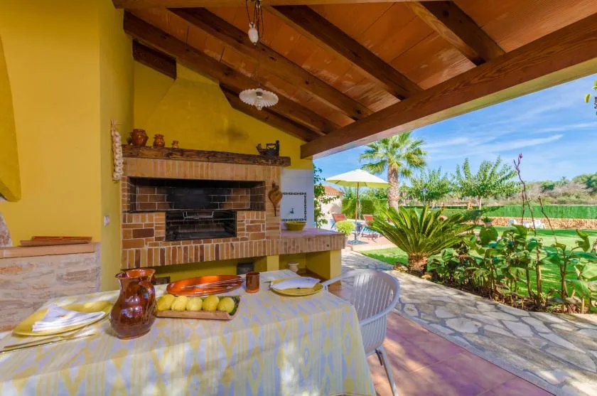 Un comedor exterior cubierto tiene una mesa junto a una barbacoa de ladrillo y un jardín.