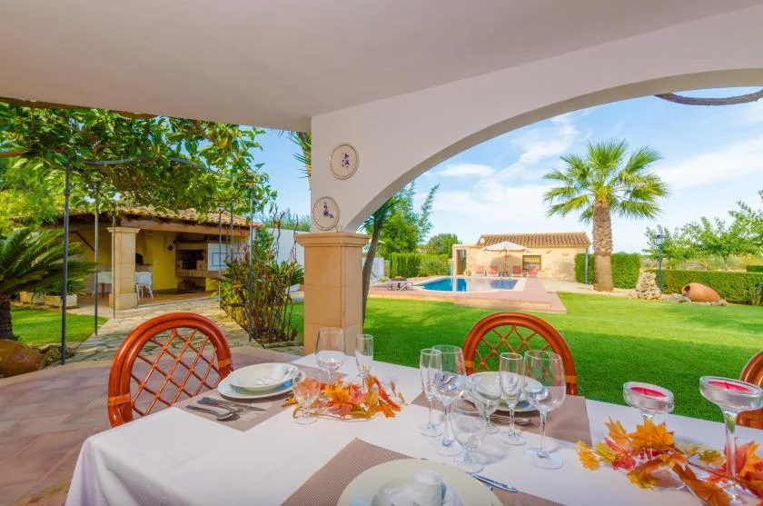 Una zona de comedor exterior cubierta da a un césped verde, una palmera y una piscina.