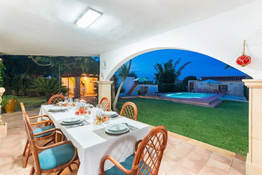 Una zona de comedor exterior cubierta da a una piscina y un césped al anochecer.