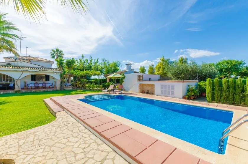 Una piscina junto a un patio pavimentado y césped verde bajo un cielo soleado.