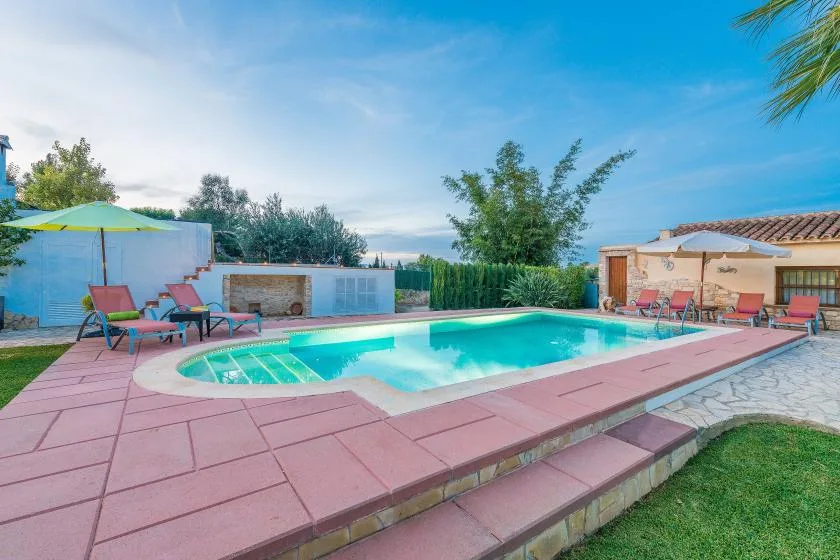 Una piscina al aire libre está rodeada por una terraza pavimentada, asientos y vegetación.