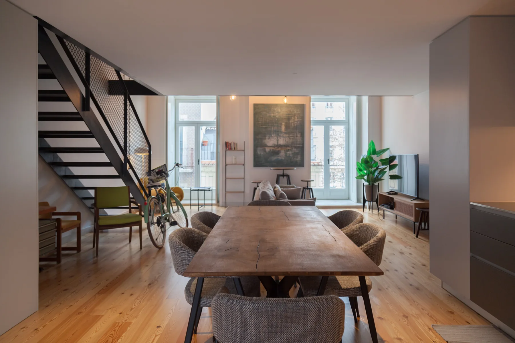 A dining table sits in a bright open-plan room with a staircase and large windows.