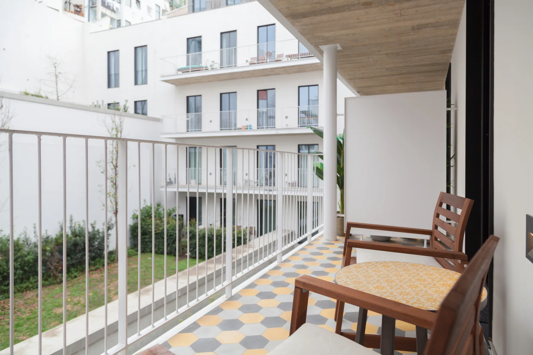 Un balcón con suelo de baldosas y asientos de madera da a un patio y un edificio cercano.