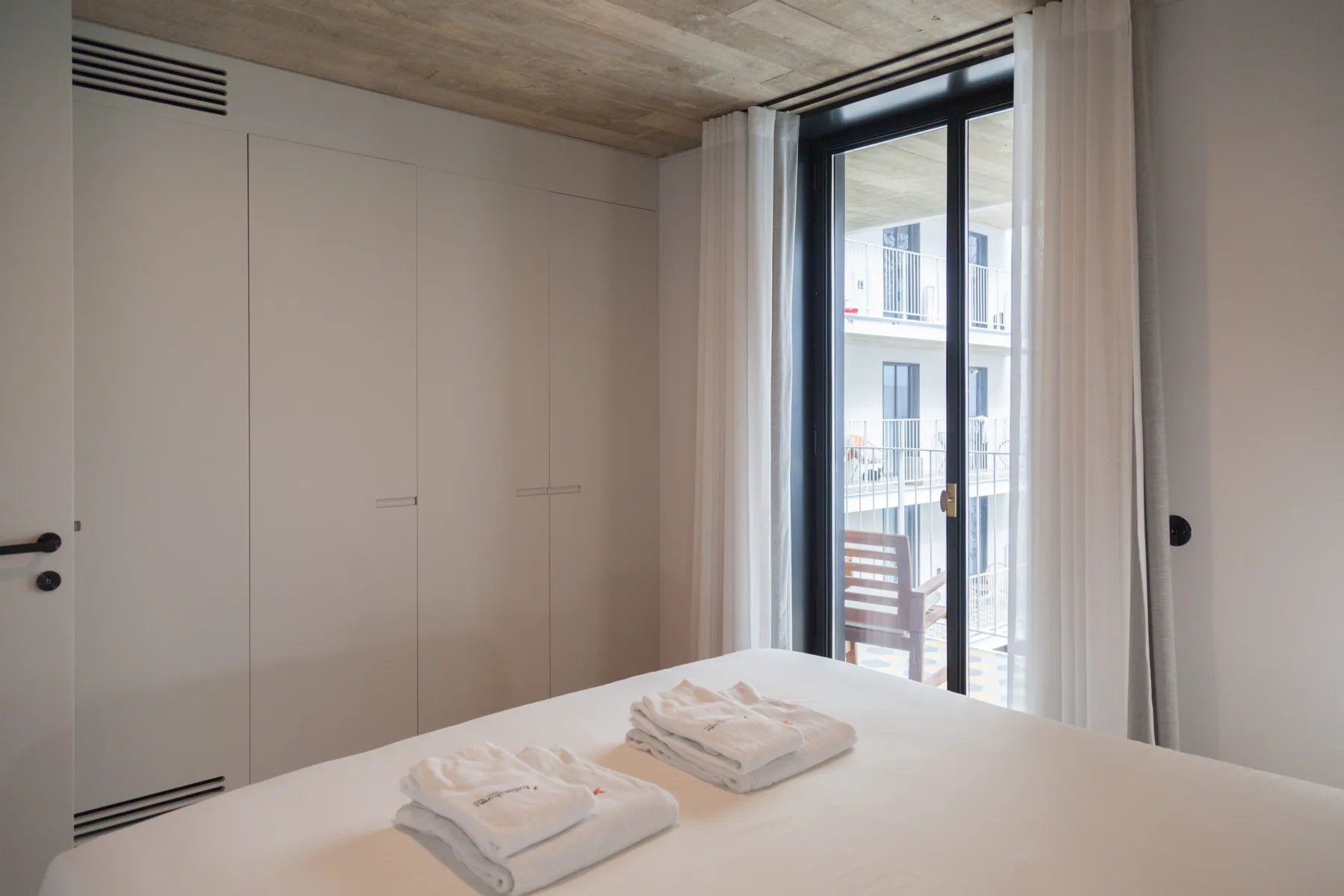 A bedroom with a bed beside large glass doors and white curtains.