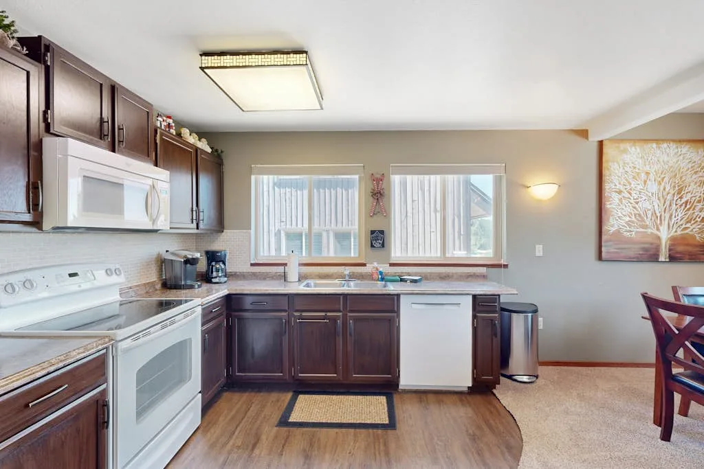 A kitchen with dark cabinets, appliances, and two windows above the counter.