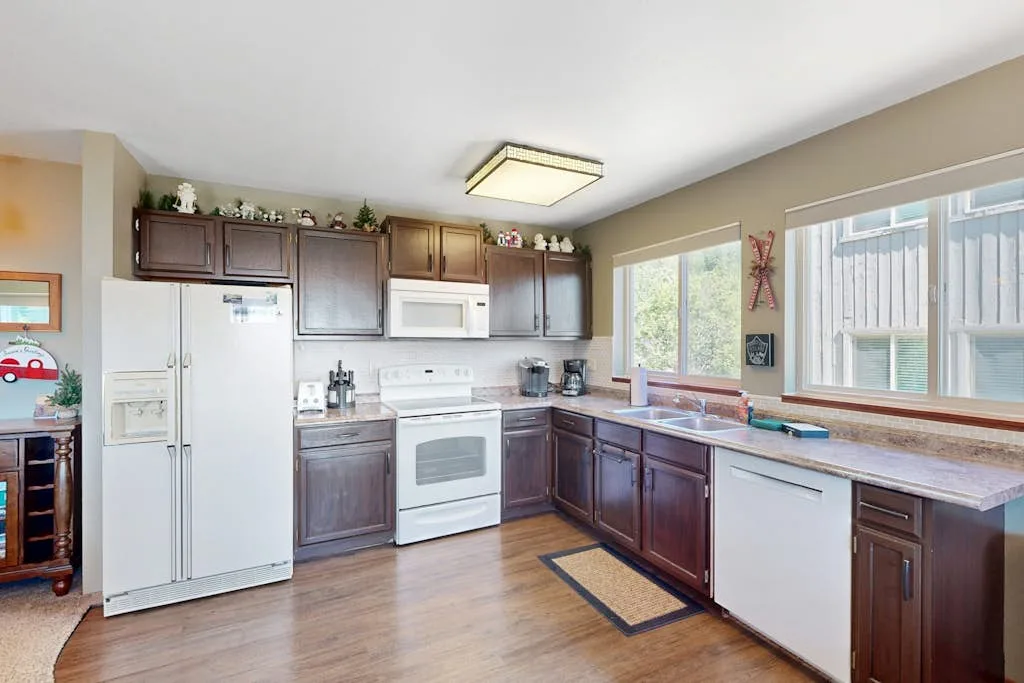 A kitchen with dark cabinets, white appliances, and windows above the counter.