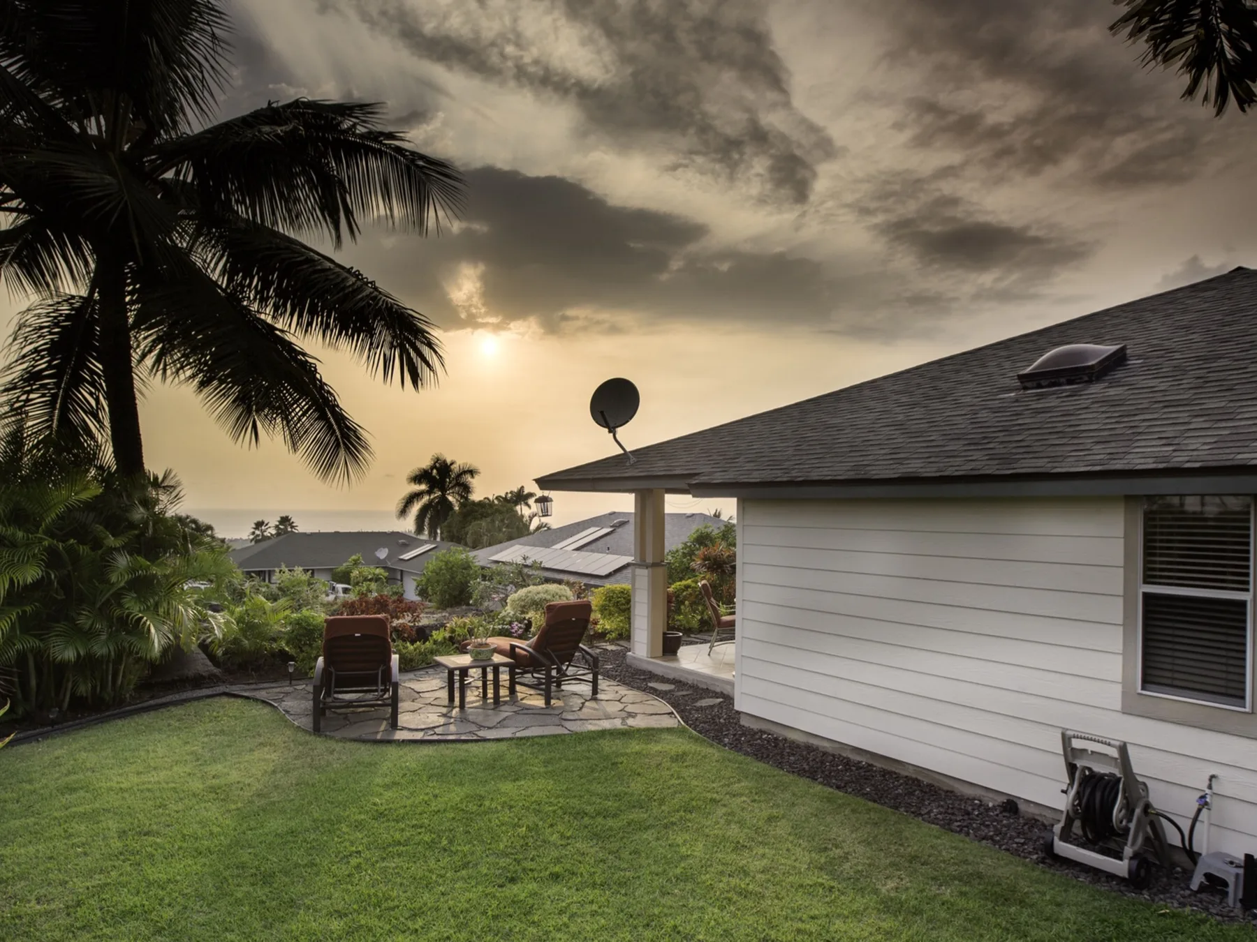 Un patio ajardinado con asientos al aire libre junto a una casa de revestimiento blanco, bajo un cielo nublado al atardecer.