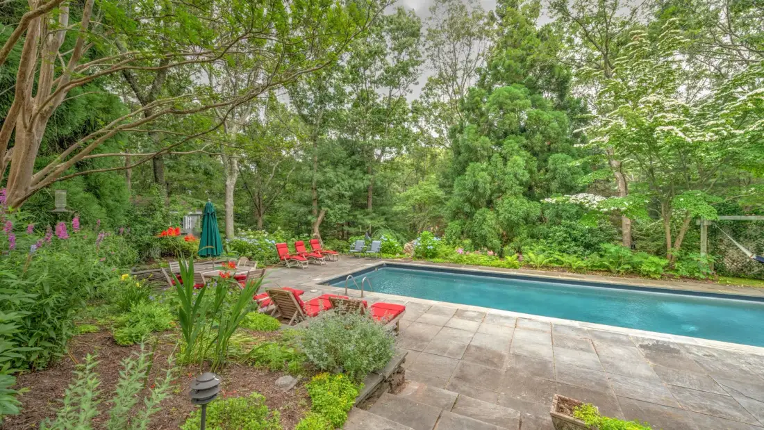 An outdoor swimming pool is surrounded by trees, garden beds, and patio seating.