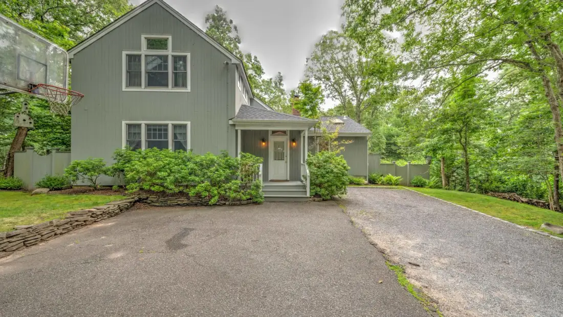 A gray house stands beside a paved driveway, surrounded by trees and shrubs.