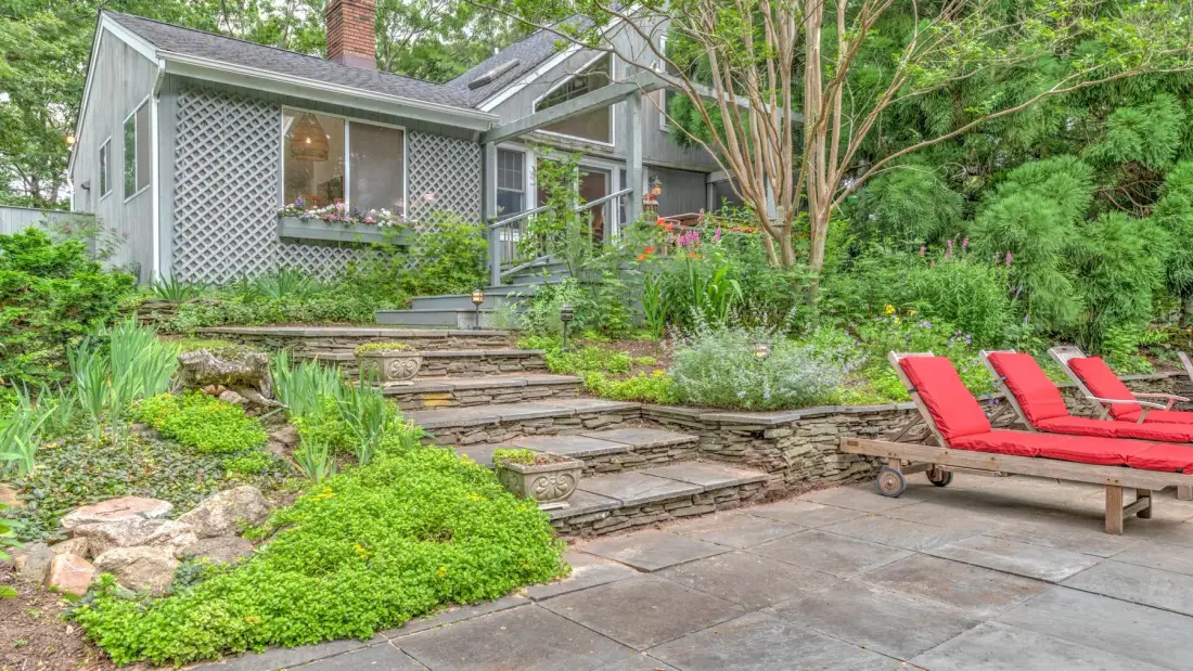 A garden patio with stone steps leads toward a house surrounded by greenery.