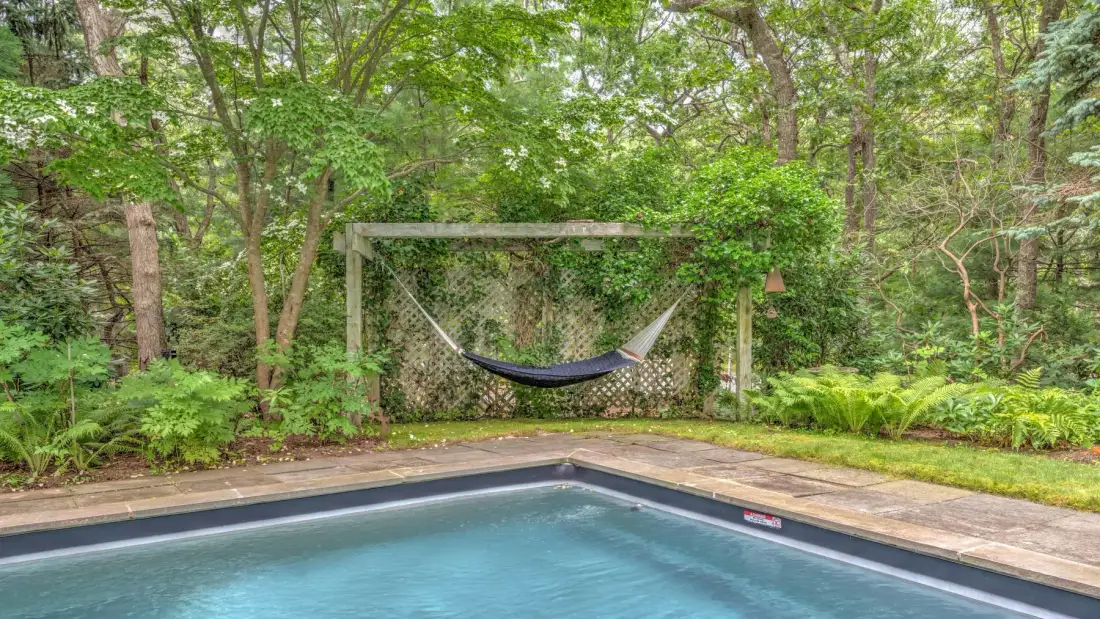 An outdoor swimming pool sits beside a wooded garden with a hammock.