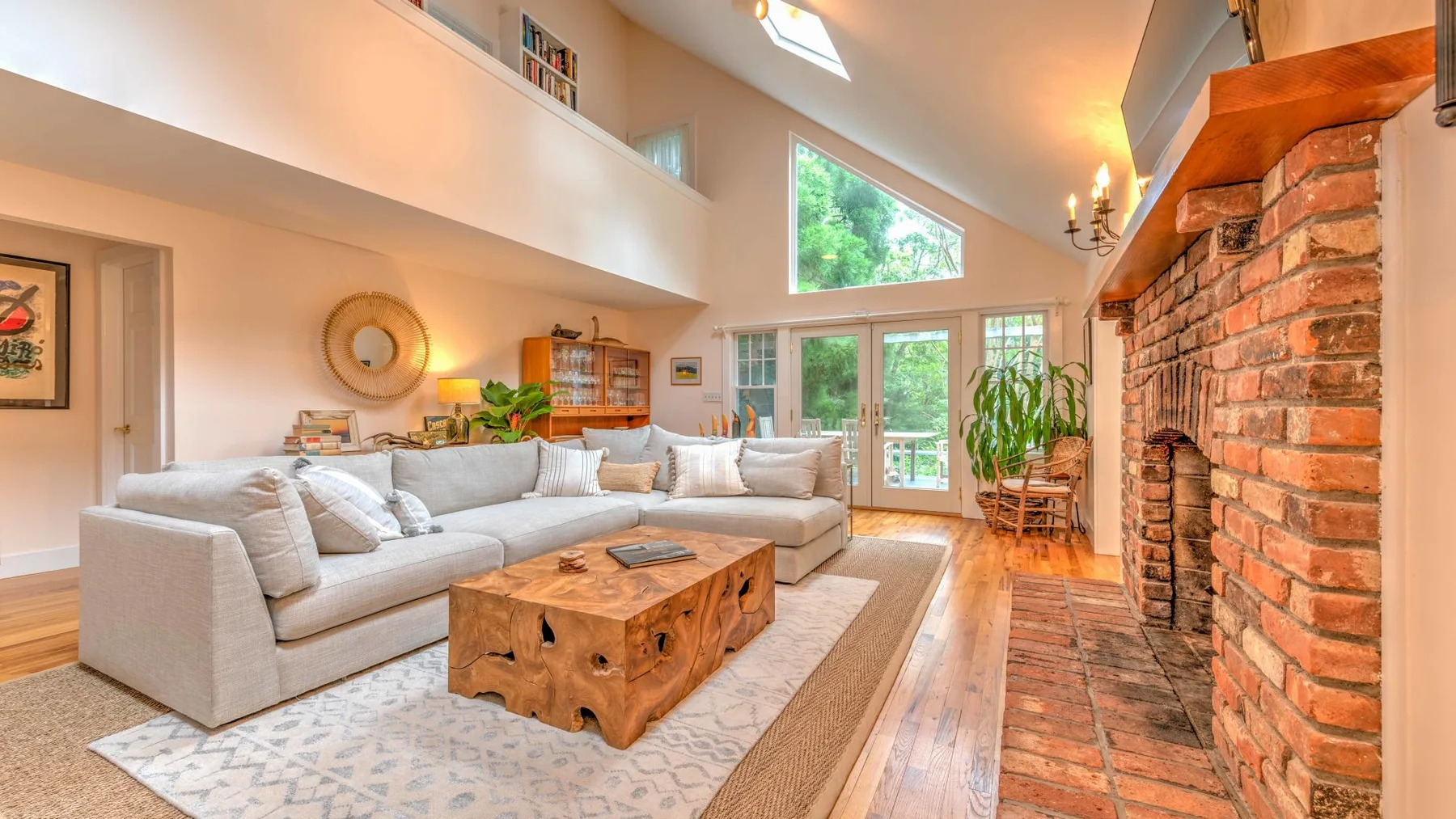 A bright living room with seating, a wooden coffee table, and a large brick fireplace.