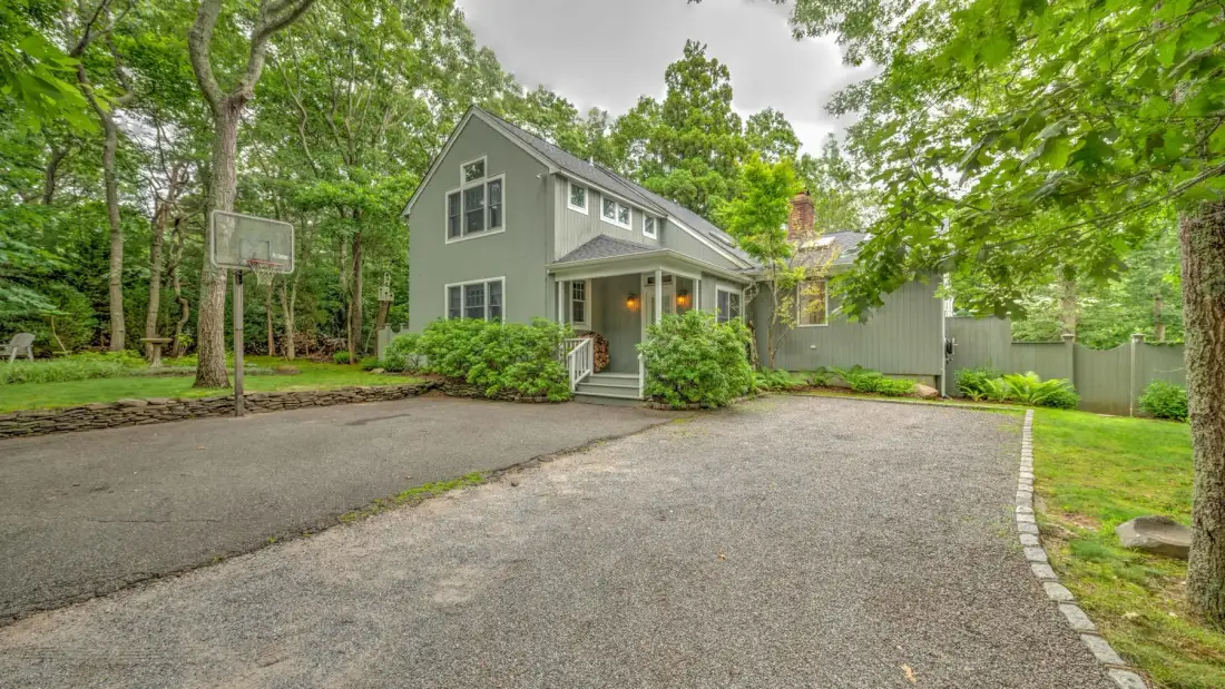 A gray house stands beside a paved driveway, with trees surrounding the property.